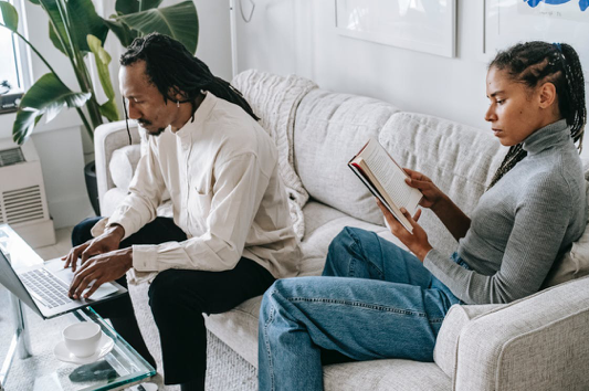 Couple sitting on a couch with Man using his laptop and woman reading a book