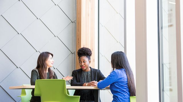 A culturally diverse group of 3 female  Royal Roads University students sit together in green chairs around a table having a conversation in one of the schools modern campus buildings.