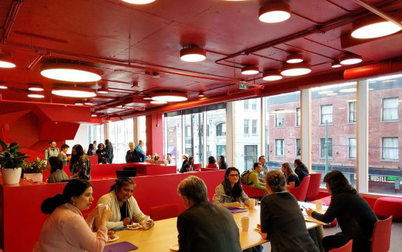 A group of students sitting around a large desk table on the campus of Adler University Vancouver in British Columbia, Canada.