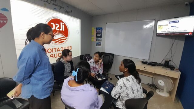 A group of women sit in a circle in a classroom, engaged in discussion. One woman stands with a phone in hand. A whiteboard and a wall-mounted screen are visible, displaying a website. The room has motivational quotes and bright colors, creating an educational and collaborative atmosphere.