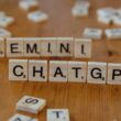Wooden Scrabble tiles spell "GEMINI" above "CHATGPT" on a wooden surface. Scattered tiles are in the background, creating a playful, creative vibe.