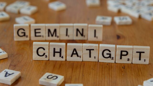 Wooden Scrabble tiles spell "GEMINI" above "CHATGPT" on a wooden surface. Scattered tiles are in the background, creating a playful, creative vibe.