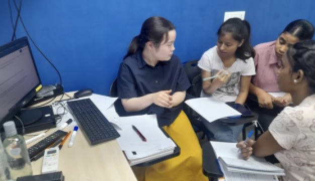 A group of students engaged in discussion around a computer, with notebooks and stationery scattered on the table in a blue-walled classroom.