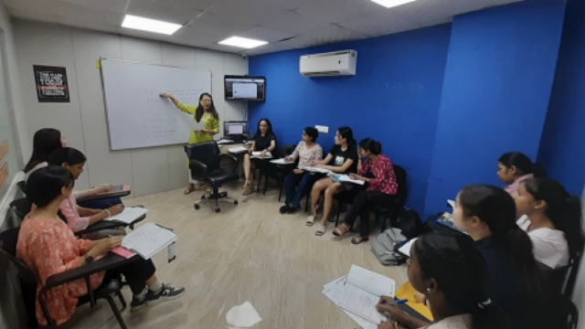 A classroom setting with a teacher instructing students seated in a semicircle, using a whiteboard and a presentation screen.