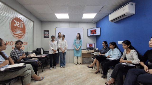 A classroom with three students standing and presenting, while others sit attentively around a table. A slide is displayed on a monitor. The atmosphere is focused and educational.